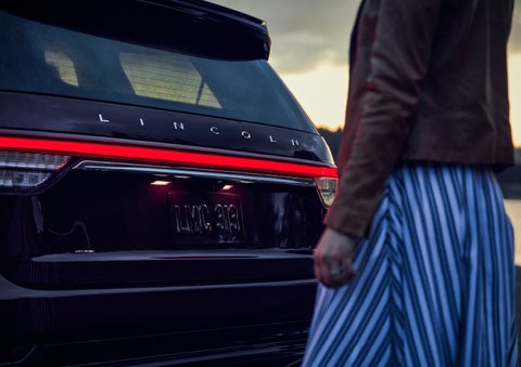 A person is shown near the rear of a 2024 Lincoln Aviator® SUV as the Lincoln Embrace illuminates the rear lights | Dave Sinclair Lincoln in St Louis MO